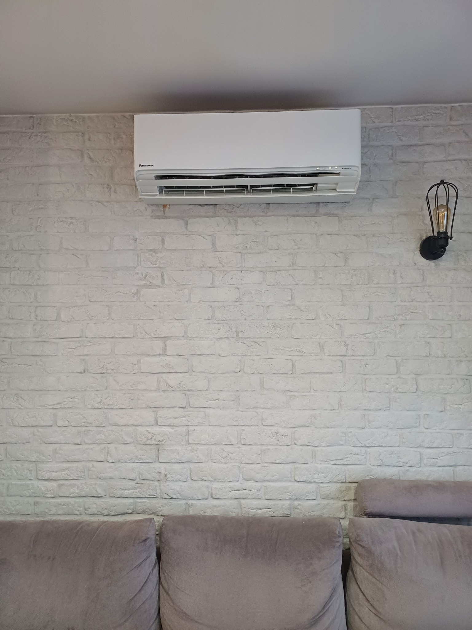 Biała klimatyzacja Panasonic zamontowana na ścianie z białej cegły, obok lampa w stylu industrialnym, widoczny fragment szarej sofy.