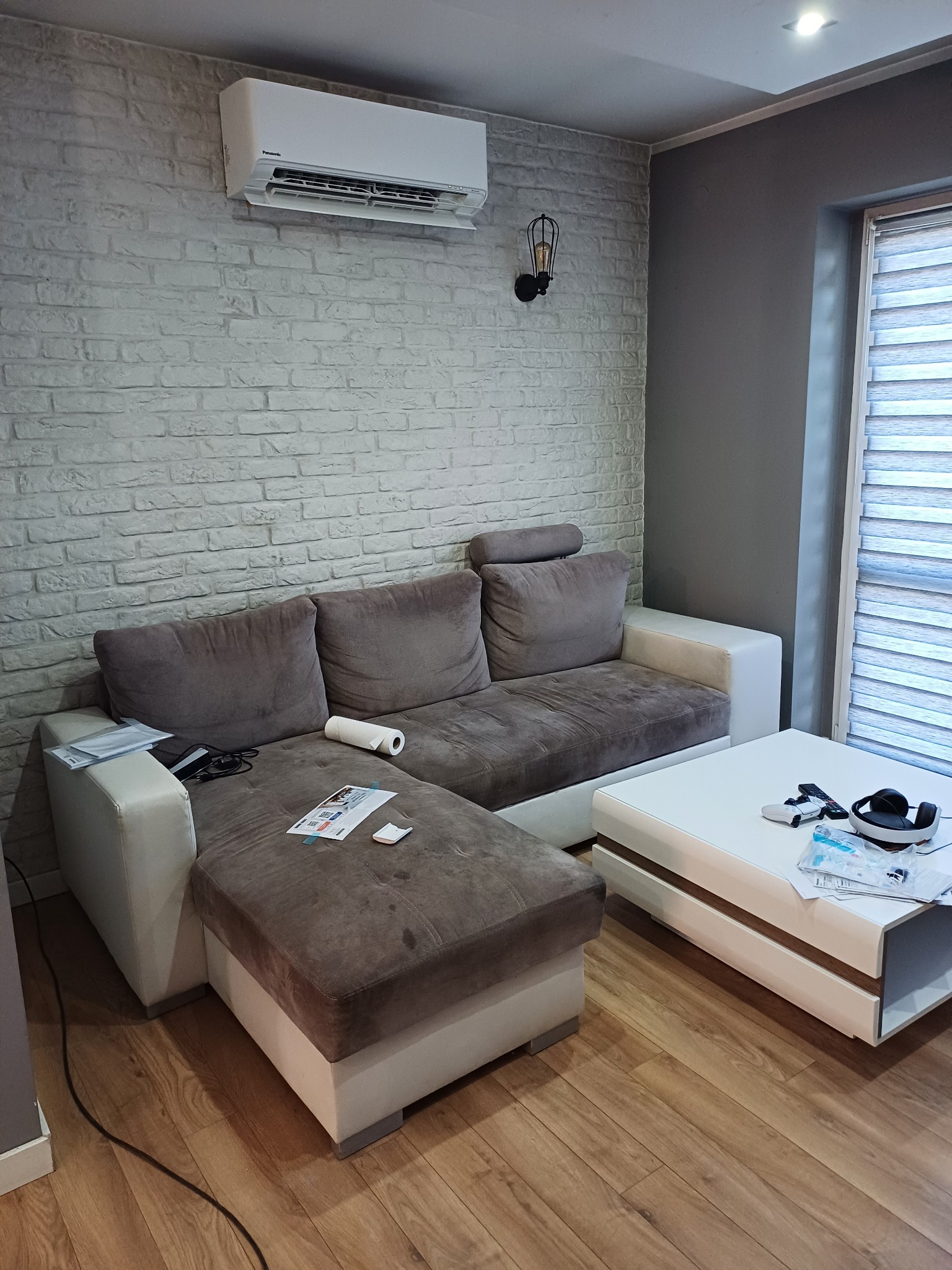 Biała klimatyzacja Panasonic zamontowana na ścianie z cegły w salonie z szarą sofą i białym stolikiem kawowym