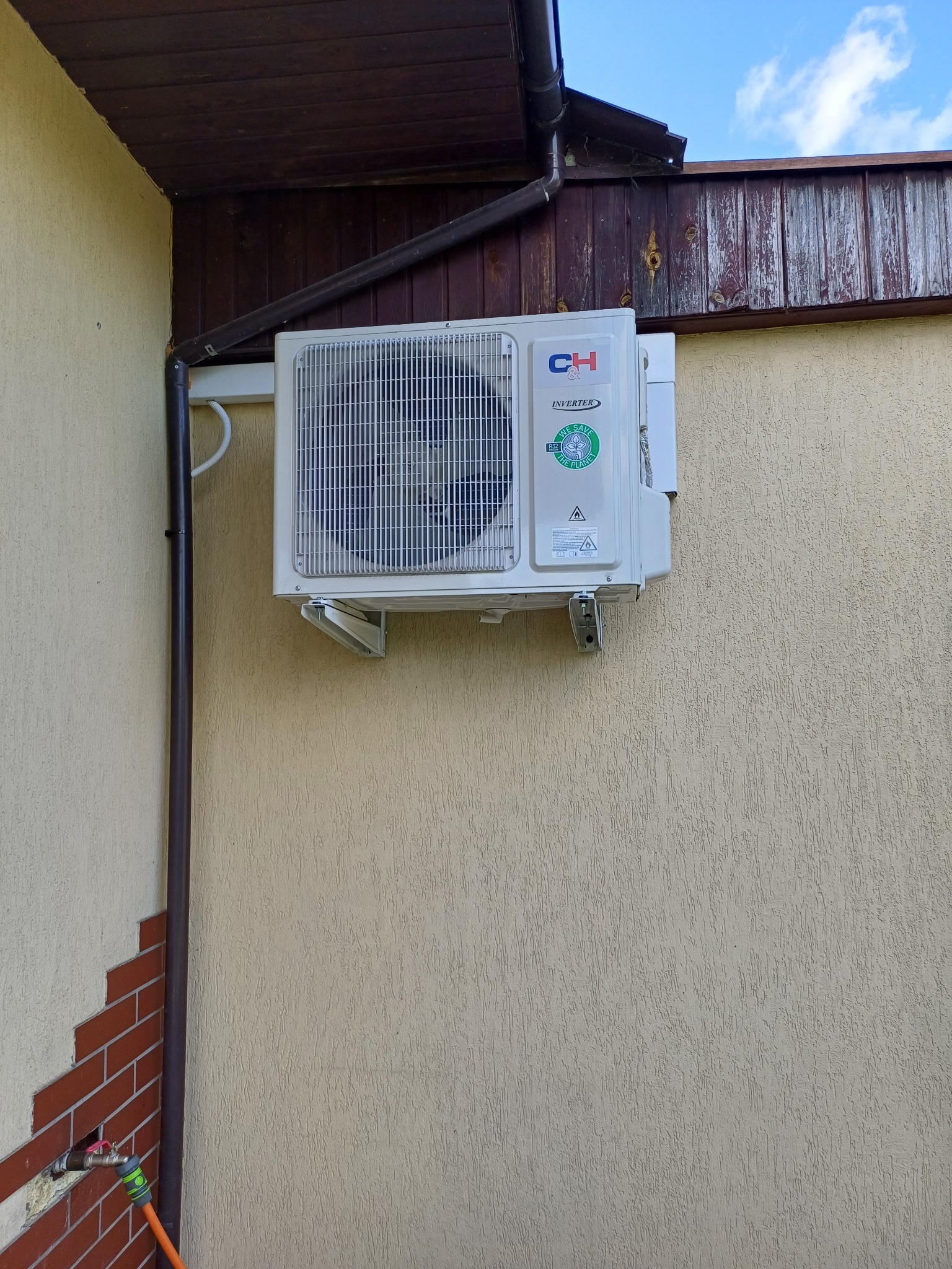 Biała jednostka zewnętrzna klimatyzatora z naklejką 'We save the planet' zamontowana na beżowej ścianie budynku z widocznym fragmentem dachu i rynny.
