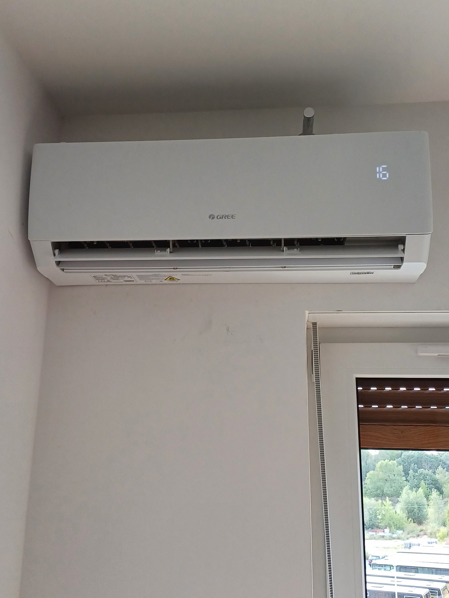Biały klimatyzator marki Gree, zamontowany na ścianie w domu w Mosinie, z wyświetlaczem wskazującym temperaturę 16 stopni Celsjusza, widok z okna na zewnątrz.