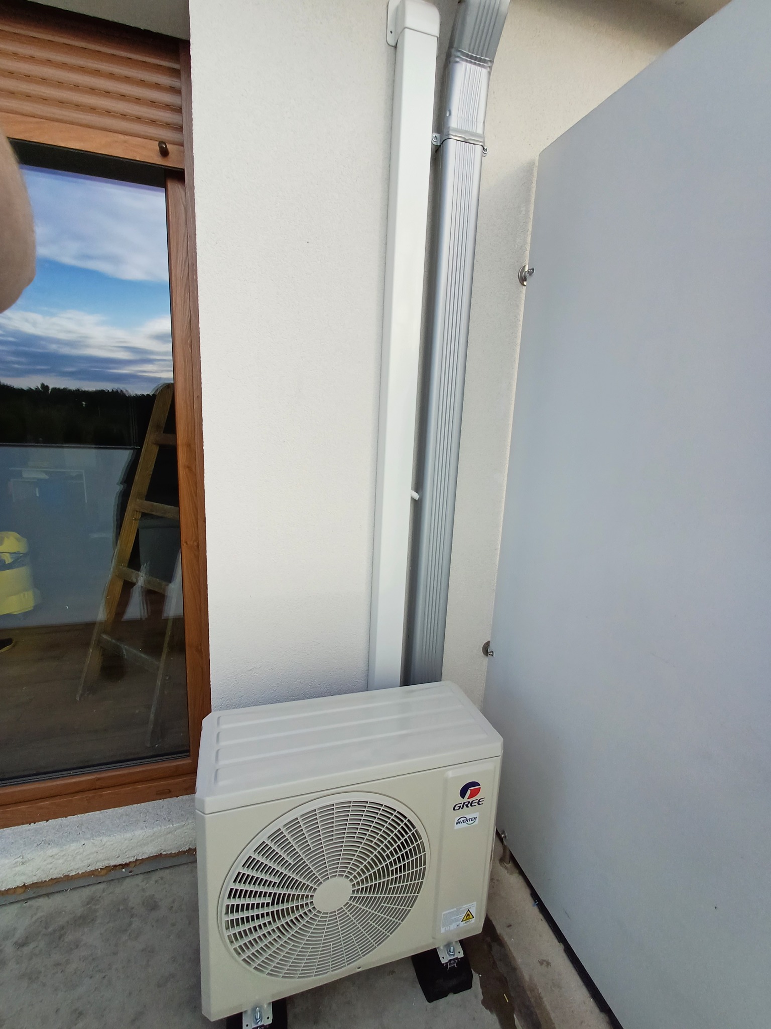 Zewnętrzna jednostka klimatyzatora marki Gree zamontowana na zewnątrz budynku, obok okna z drewnianą ramą i metalowej rynny spustowej.