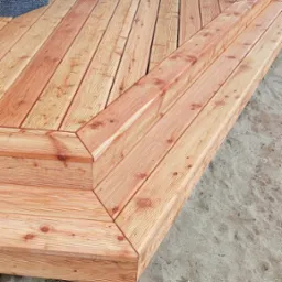 Nowoczesny taras z drewna egzotycznego z niskimi schodami, widok z góry na jasny piasek i fragment budynku z oknem.