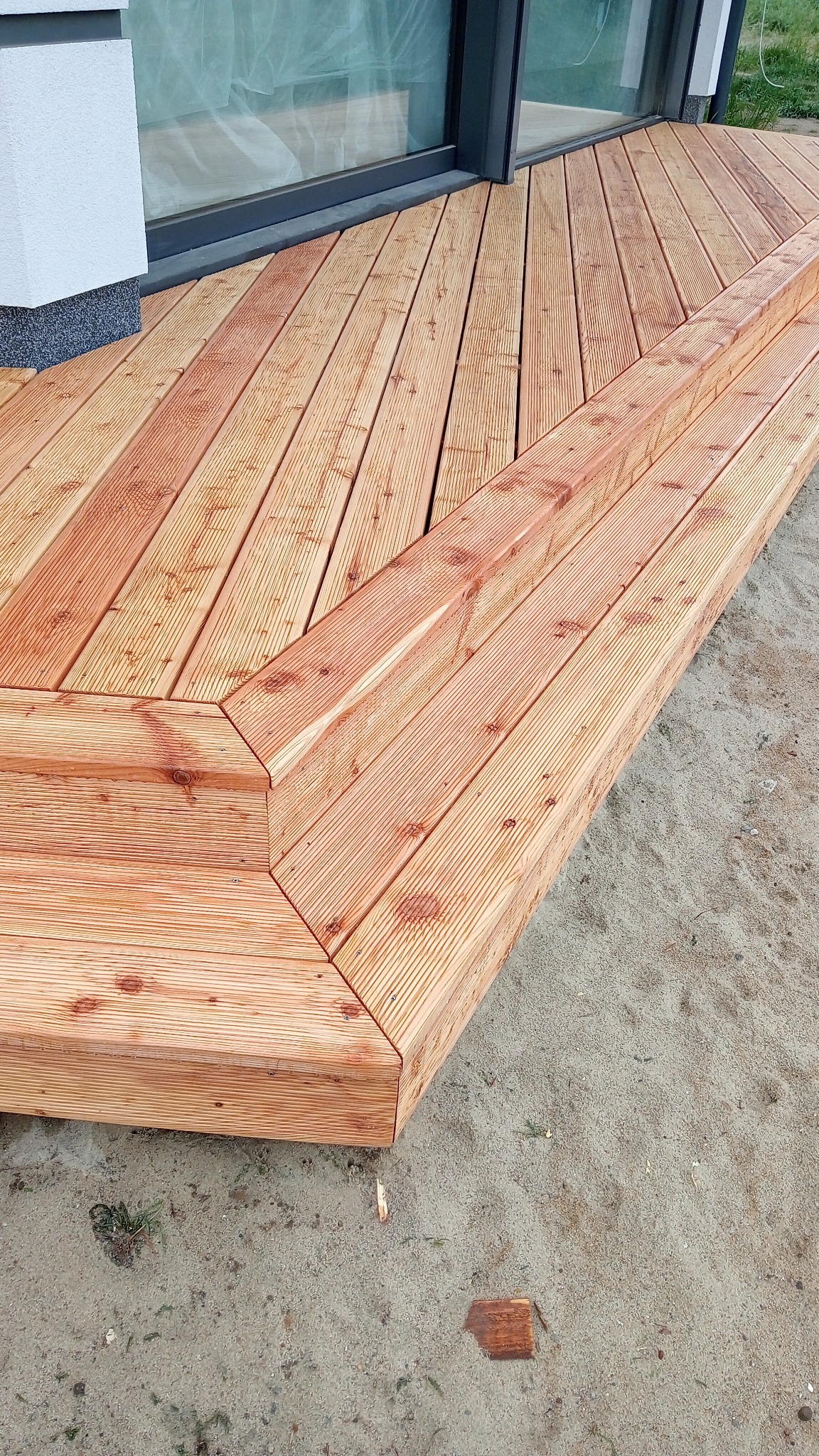 Nowoczesny taras z drewna egzotycznego z niskimi schodami, widok z góry na jasny piasek i fragment budynku z oknem.