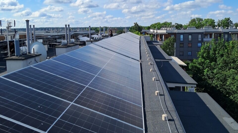 Widok z góry na instalację paneli słonecznych na płaskim dachu budynku z kominami i anteną satelitarną, w tle zabudowa miejska i błękitne niebo z chmurami.