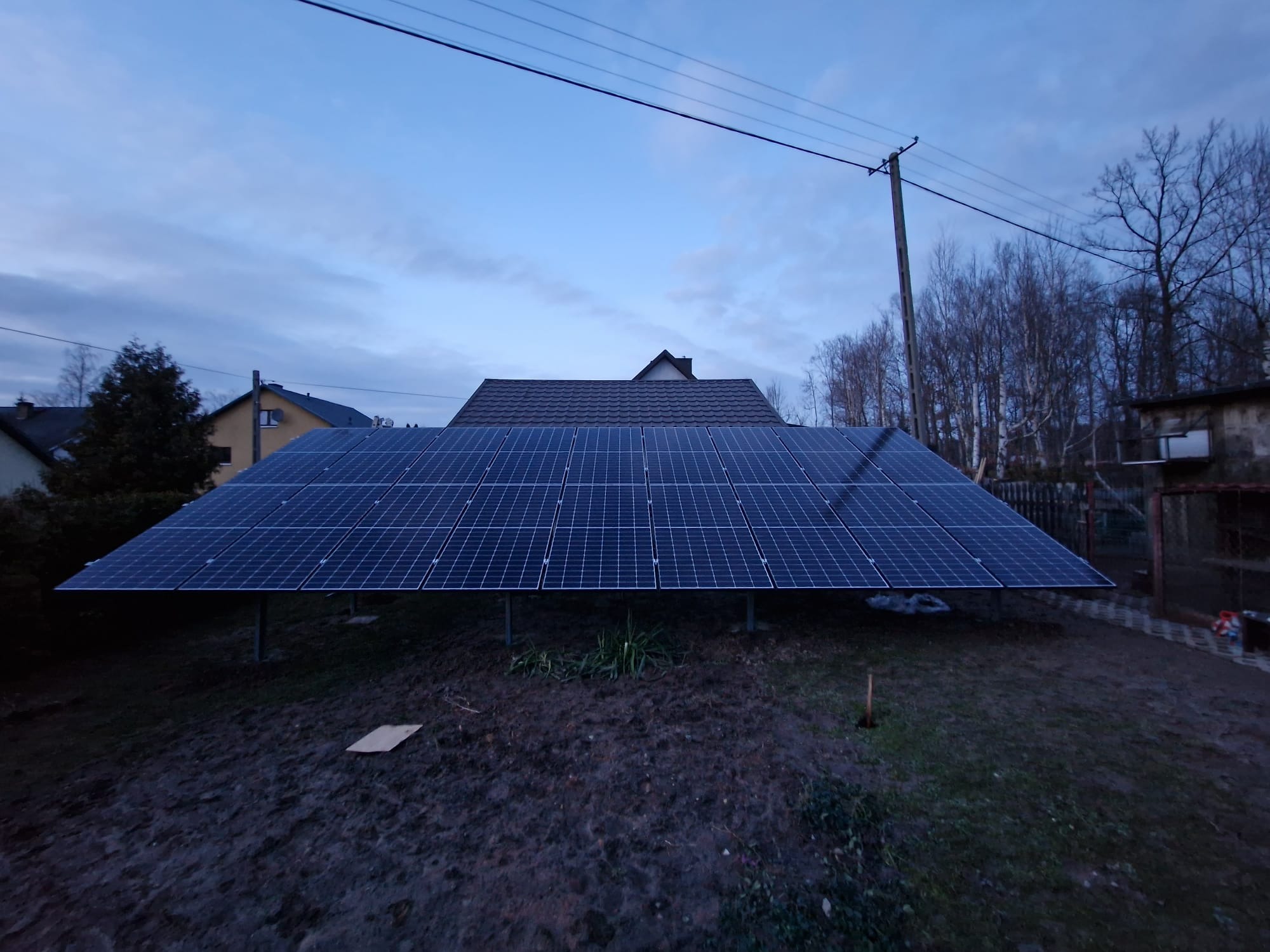 Instalacja fotowoltaiczna na metalowych podporach na działce, widok na domy i słup energetyczny w tle, pochmurne niebo.