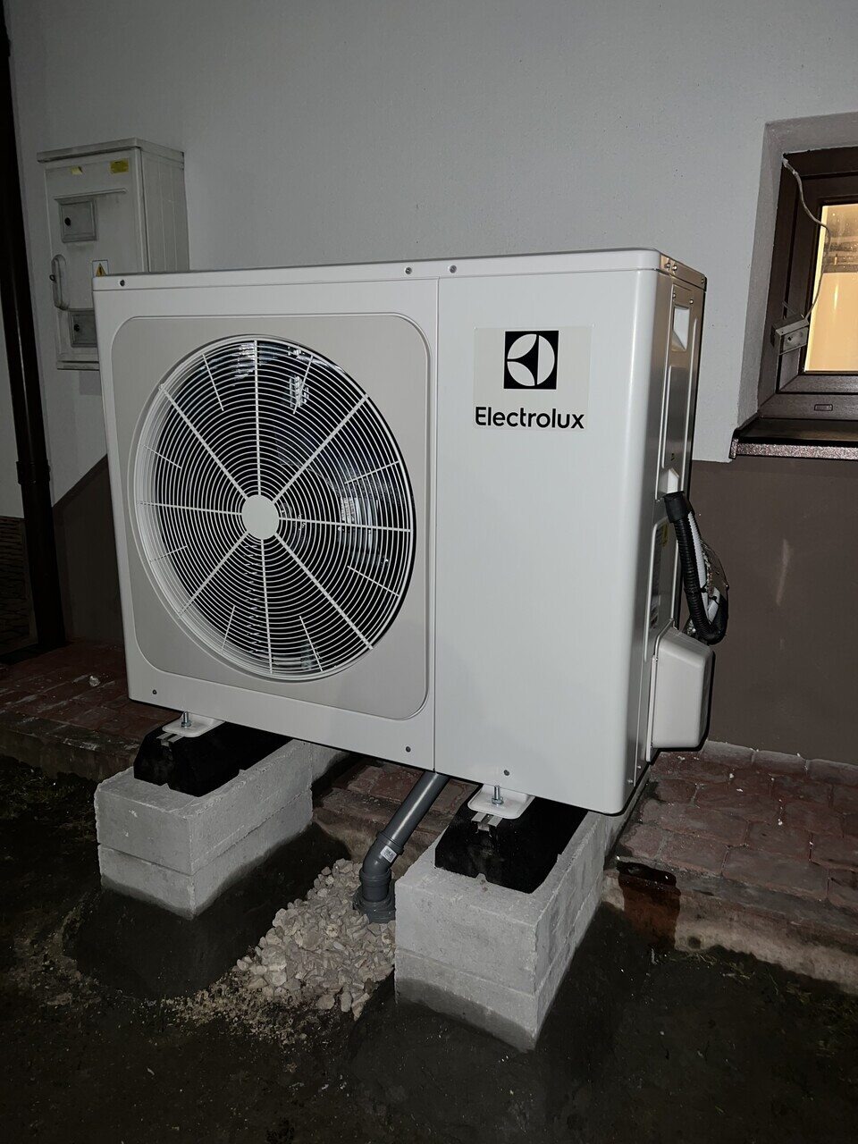 Biała, zewnętrzna jednostka pompy ciepła Electrolux, zamontowana na betonowych bloczkach z widocznym odpływem skroplin i elementami instalacji elektrycznej, umieszczona na zewnątrz budynku.