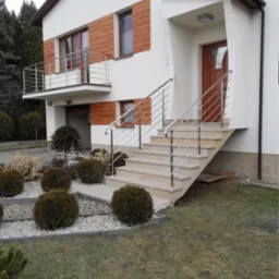 Nowoczesny dom z jasną elewacją, drewnianymi akcentami i balkonem z metalową balustradą, schody wejściowe z metalową poręczą, zadbany ogród z kulistymi krzewami i kamieniami.