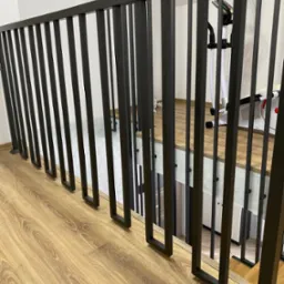 Nowoczesna balustrada wewnętrzna z czarnych, pionowych profili kwadratowych, widok z góry na drewnianą podłogę, w tle sprzęt do ćwiczeń.