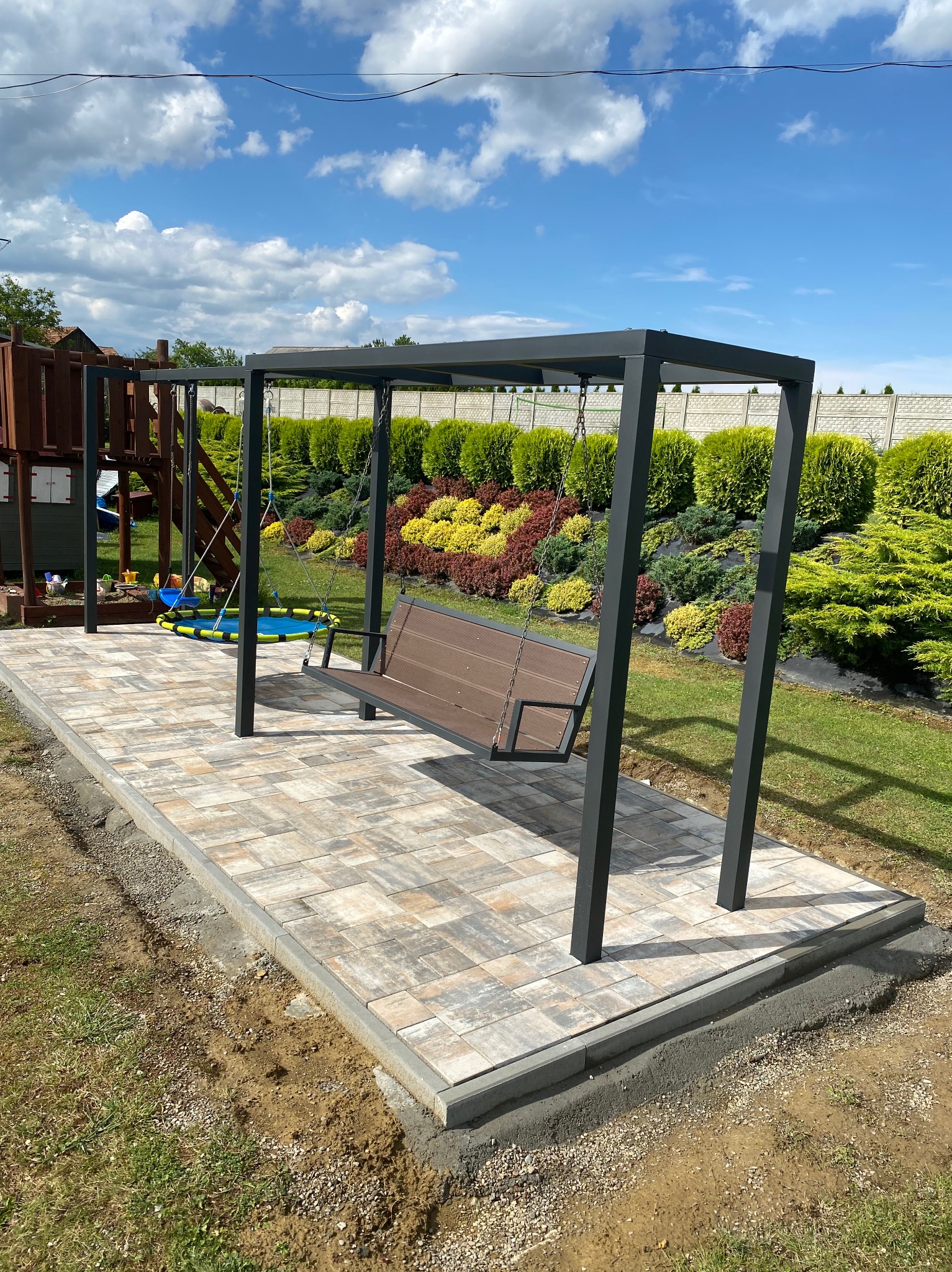 Nowoczesna huśtawka ogrodowa z metalową konstrukcją i drewnianym siedziskiem, umieszczona na wybrukowanym placu, w tle zielony ogród z tujami i plac zabaw.