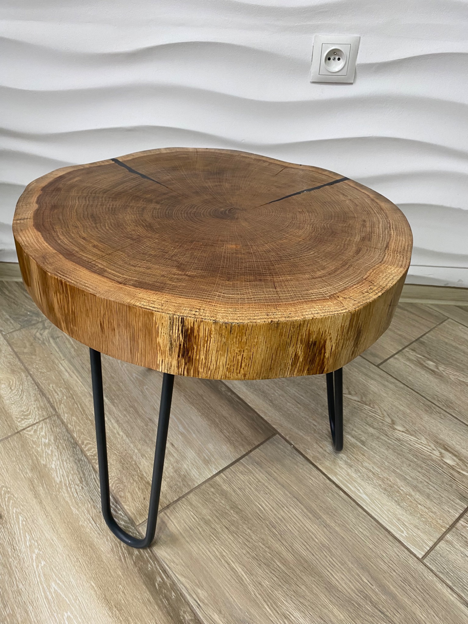 Stolik kawowy z blatem z plastra drewna z widocznymi słojami i pęknięciami, osadzony na trzech czarnych, metalowych nogach typu hairpin, na tle białej, falistej ściany i podłogi imitującej drewno.