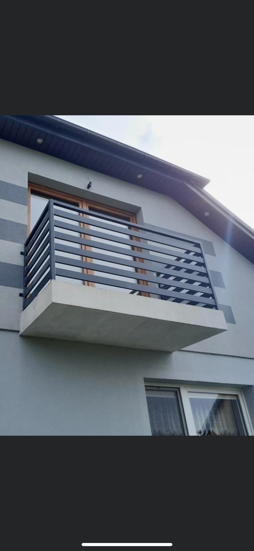 Nowoczesny balkon z czarną, poziomą balustradą na tle szarej elewacji domu z drewnianą ramą okienną.