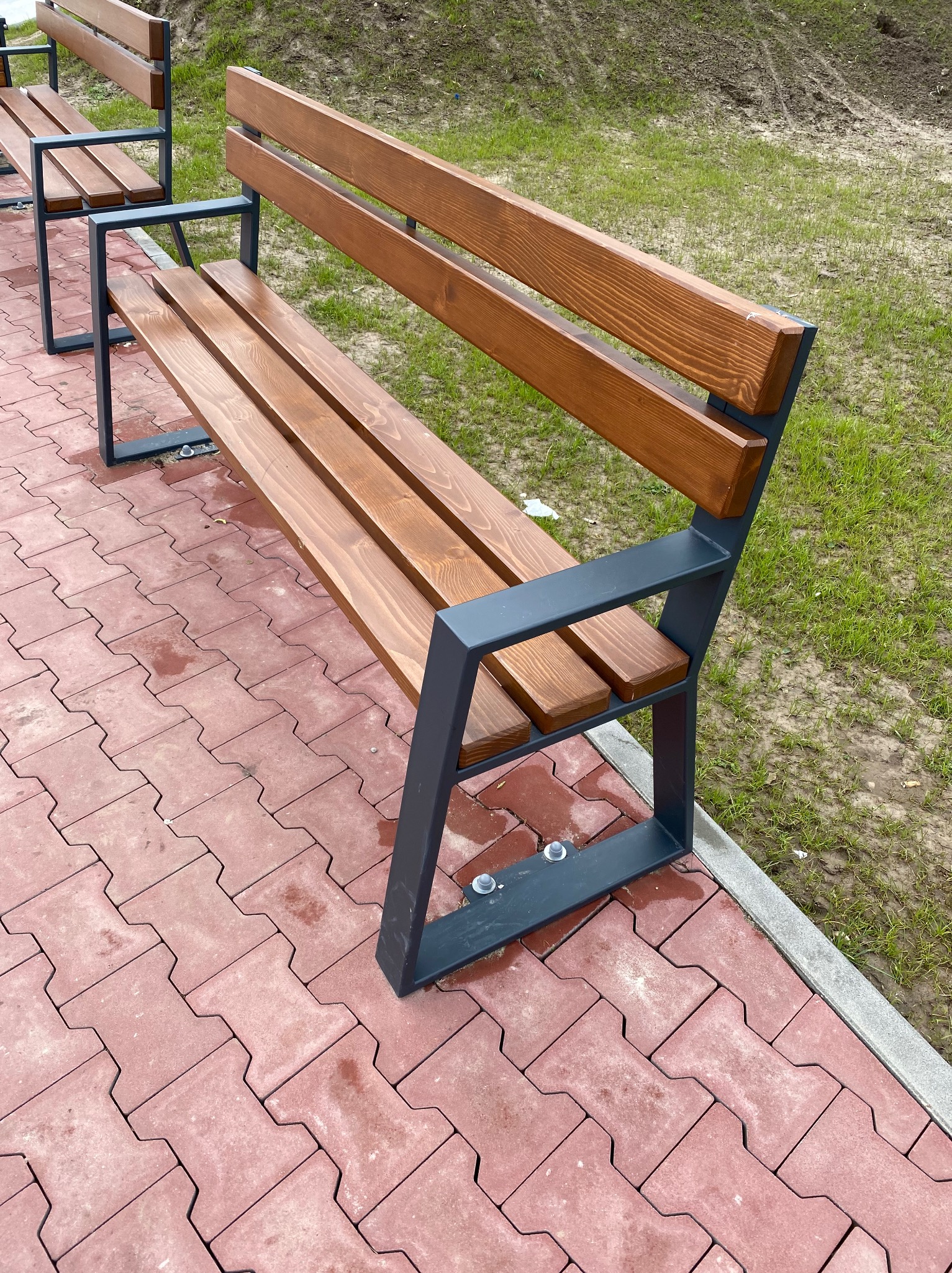 Nowoczesna ławka parkowa z drewnianym siedziskiem i szarymi metalowymi elementami, ustawiona na czerwonej kostce brukowej obok trawnika.
