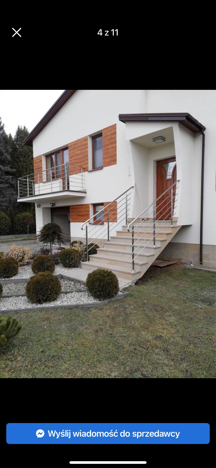 Nowoczesny dom z jasną elewacją, drewnianymi akcentami i balkonem z metalową balustradą, schody wejściowe z metalową poręczą, zadbany ogród z kulistymi krzewami i kamieniami.