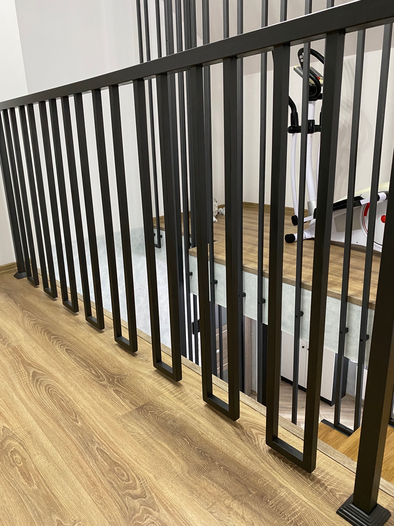 Nowoczesna balustrada wewnętrzna z czarnych, pionowych profili kwadratowych, widok z góry na drewnianą podłogę, w tle sprzęt do ćwiczeń.