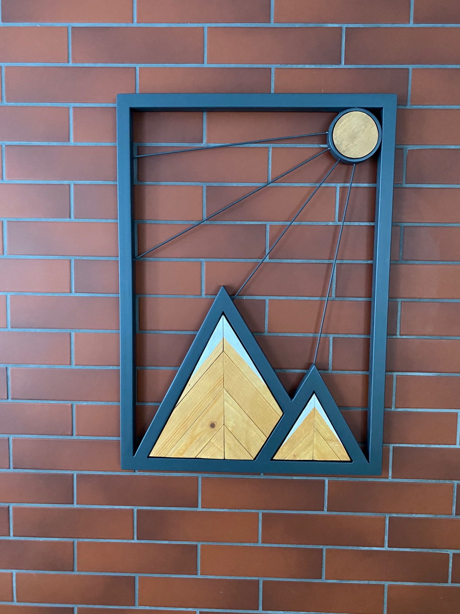 Dekoracja ścienna: metalowa rama z drewnianymi górami i słońcem na tle ceglanej ściany.