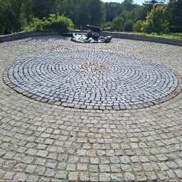Ogrody Bonsai Damian Kopiec - Okrągła nawierzchnia z kostki granitowej w parku, z centralnym wzorem w jaśniejszym odcieniu. W tle zieleń drzew i krzewów, tworząca naturalne tło dla kamiennej kompozycji.