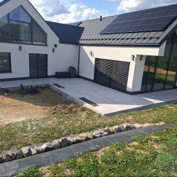 Ogrody Bonsai Damian Kopiec - Nowoczesny taras z jasnych płyt przy białym domu z panelami słonecznymi. Częściowo wykończony ogród z obrzeżami z kamieni i czarnej agrowłókniny.