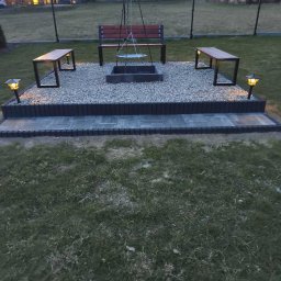 Ogrody Bonsai Damian Kopiec - Ogród z miejscem na ognisko: kamienny krąg, ławki z drewna i metalu, oświetlenie. Całość otoczona trawnikiem i ogrodzeniem z siatki.