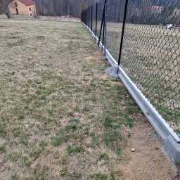 Ogrody Bonsai Damian Kopiec - Długi, czarny płot z siatki, osadzony na betonowej podmurówce, rozciąga się wzdłuż działki z widokiem na dom i zalesione wzgórza w tle. Ujęcie z bliska, z perspektywy ziemi.