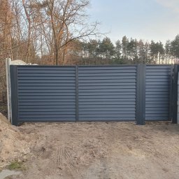 Ogrody Bonsai Damian Kopiec - Nowoczesne, poziome ogrodzenie panelowe w kolorze grafitowym, z betonowymi słupkami, na tle drzew i nieba. Widoczne ślady opon na piasku.