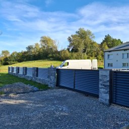Ogrody Bonsai Damian Kopiec - Nowoczesne ogrodzenie panelowe z przesuwną bramą, szare panele i kamienne słupki, na tle zielonego trawnika i błękitnego nieba. Widoczny biały bus.