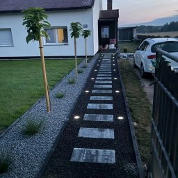Ogrody Bonsai Damian Kopiec - Oświetlona ścieżka z płyt prowadząca do domu, obok trawnik i młode drzewka. Dekoracyjny żwir, lampy wbudowane w ścieżkę. Widok na dom i fragment samochodu.