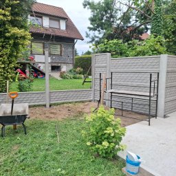 Ogrody Bonsai Damian Kopiec - Szare, prefabrykowane panele ogrodzeniowe z wzorem cegły, częściowo zamontowane. Widoczna taczka z łopatą, świeżo przekopana ziemia, trawnik i krzew hortensji. W tle dom.