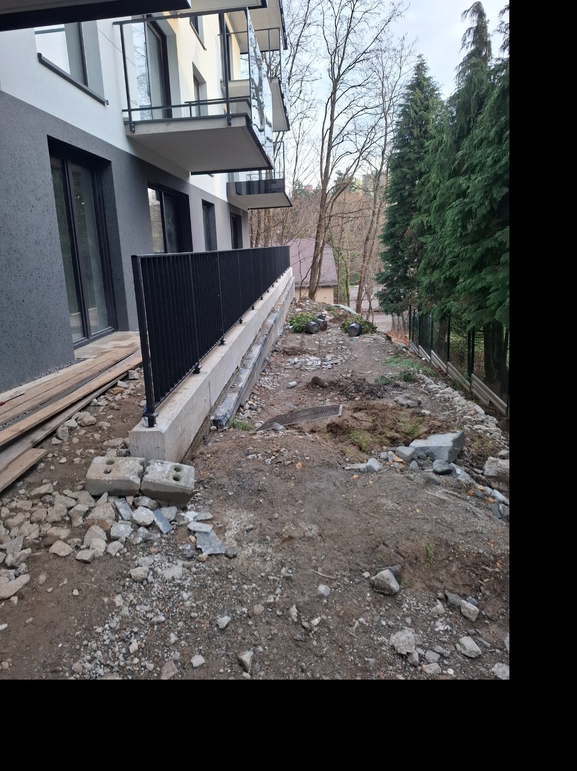 Montaż czarnej, metalowej barierki przy betonowym murku oporowym na nierównym terenie. W tle budynek z balkonami i drzewa iglaste.