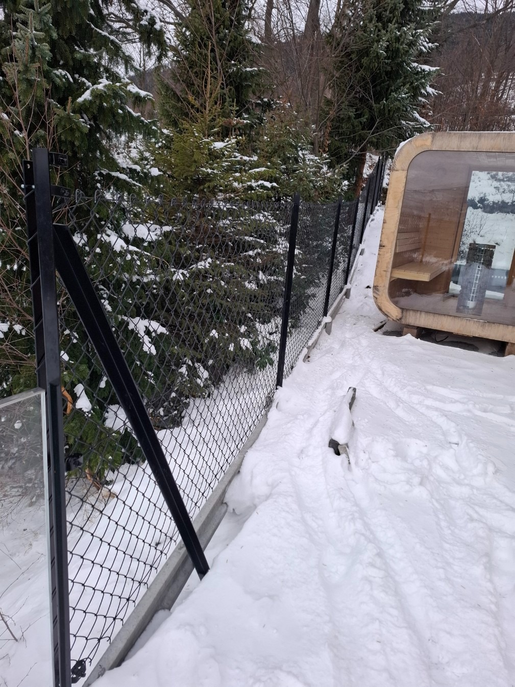 Czarny, metalowy płot z siatki na betonowej podmurówce, otoczony śniegiem i zielonymi drzewami iglastymi. W tle widoczna drewniana konstrukcja.