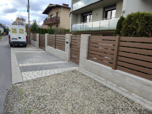 Nowoczesne ogrodzenie z poziomych paneli w kolorze brązowym, osadzone na niskim murku z jasnych bloczków. Podjazd wyłożony kostką brukową, w tle budynek mieszkalny.