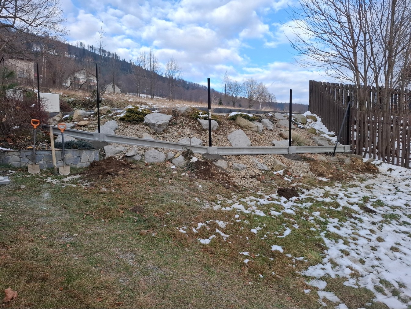 Wykop pod fundament ogrodzenia z betonową podmurówką, widoczne czarne słupki i częściowo drewniane ogrodzenie. W tle wzgórze i błękitne niebo z chmurami.