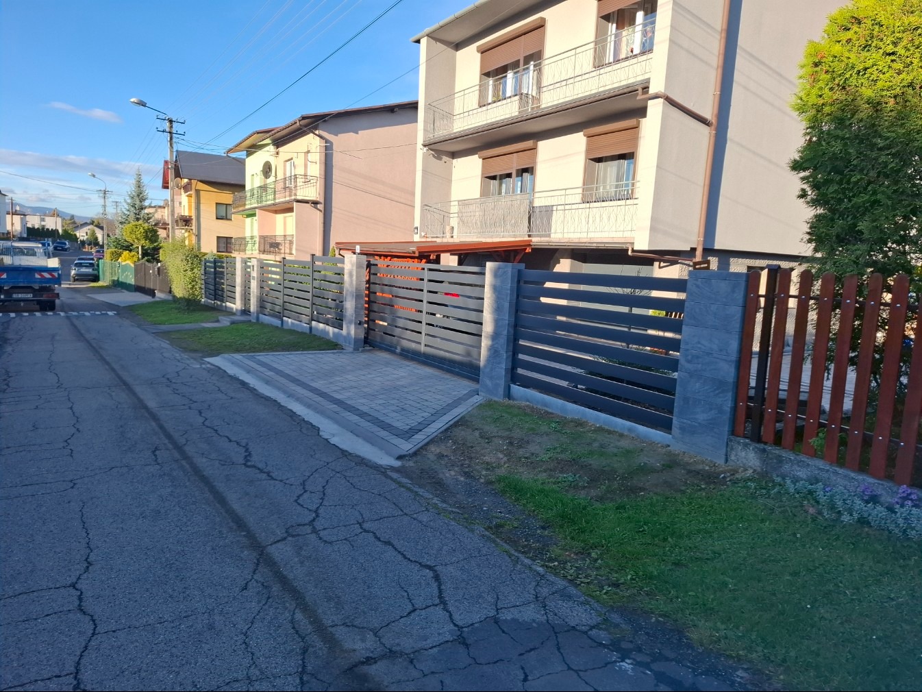 Nowoczesne, poziome ogrodzenie panelowe w kolorze antracytowym z szarymi słupkami, oddzielające posesję od ulicy. Widok na domy w tle i fragment drewnianego ogrodzenia.