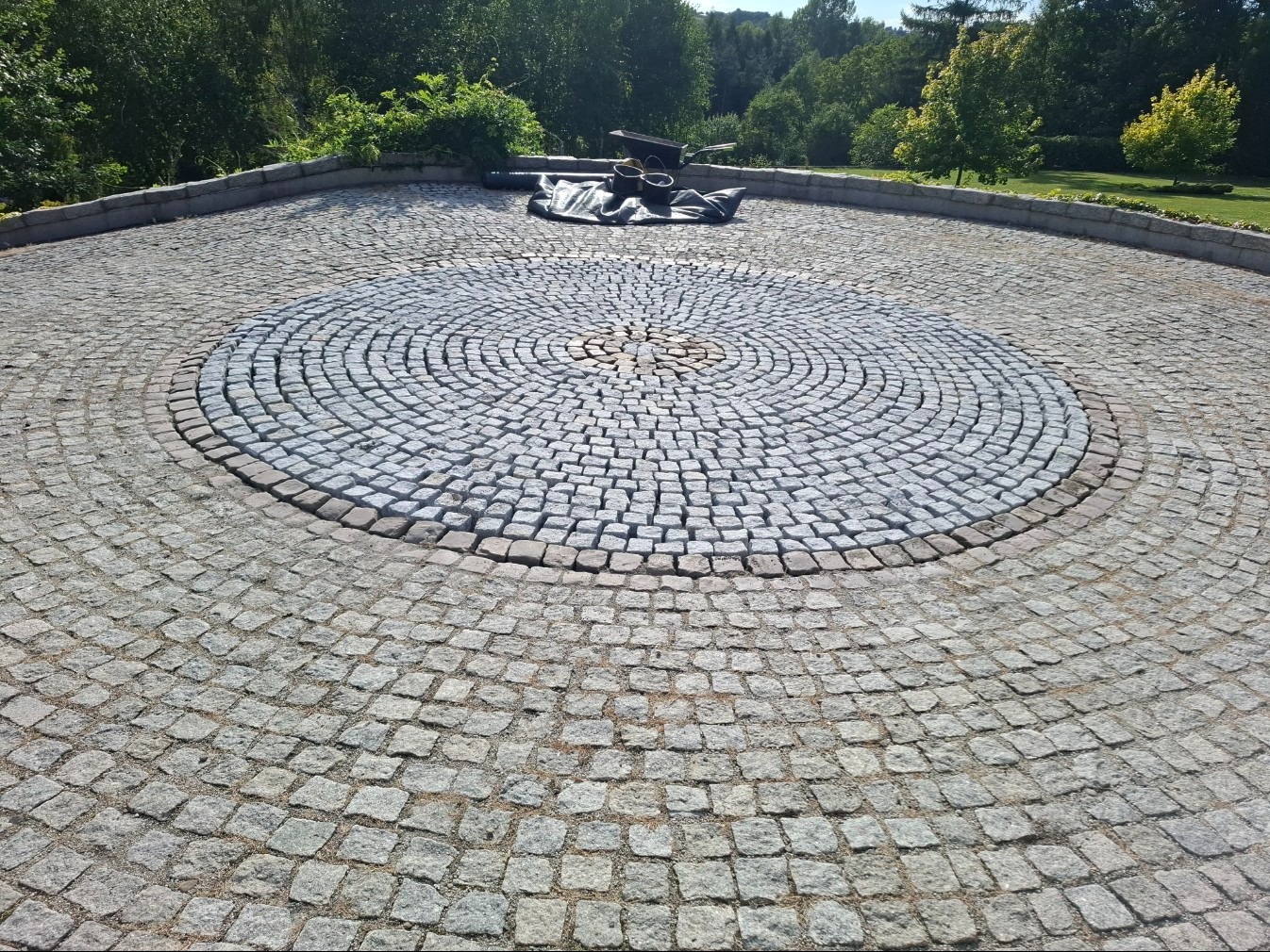 Okrągła nawierzchnia z kostki granitowej w parku, z centralnym wzorem w jaśniejszym odcieniu. W tle zieleń drzew i krzewów, tworząca naturalne tło dla kamiennej kompozycji.