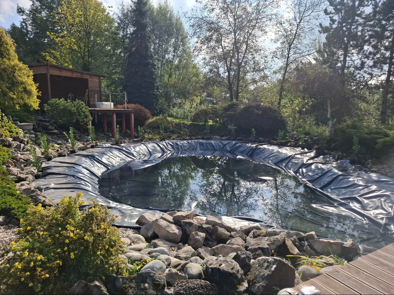 Folia w wykopanym stawie ogrodowym otoczonym kamieniami i zielenią, z drewnianą altaną w tle. Widok na częściowo wypełniony wodą zbiornik w słoneczny dzień.