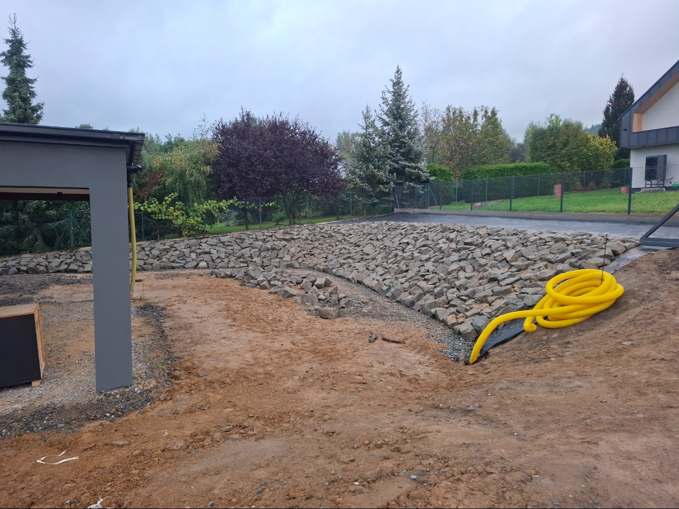 Ukończony fragment ogrodu z kamiennym murem oporowym i przygotowanym terenem pod nasadzenia. Żółty wąż ogrodowy leży na ziemi. Widok na dom i zieleń w tle.