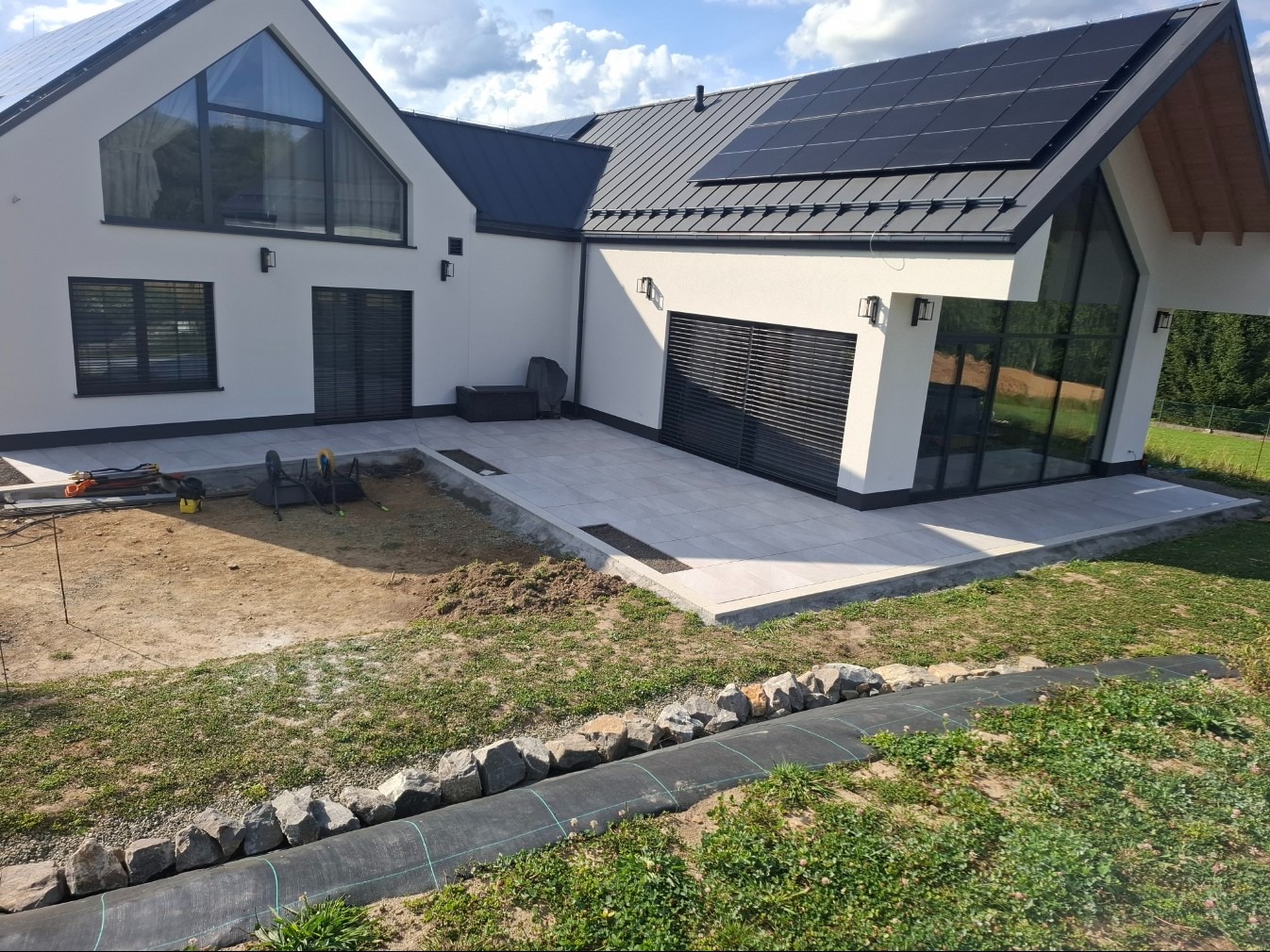 Nowoczesny taras z jasnych płyt przy białym domu z panelami słonecznymi. Częściowo wykończony ogród z obrzeżami z kamieni i czarnej agrowłókniny.