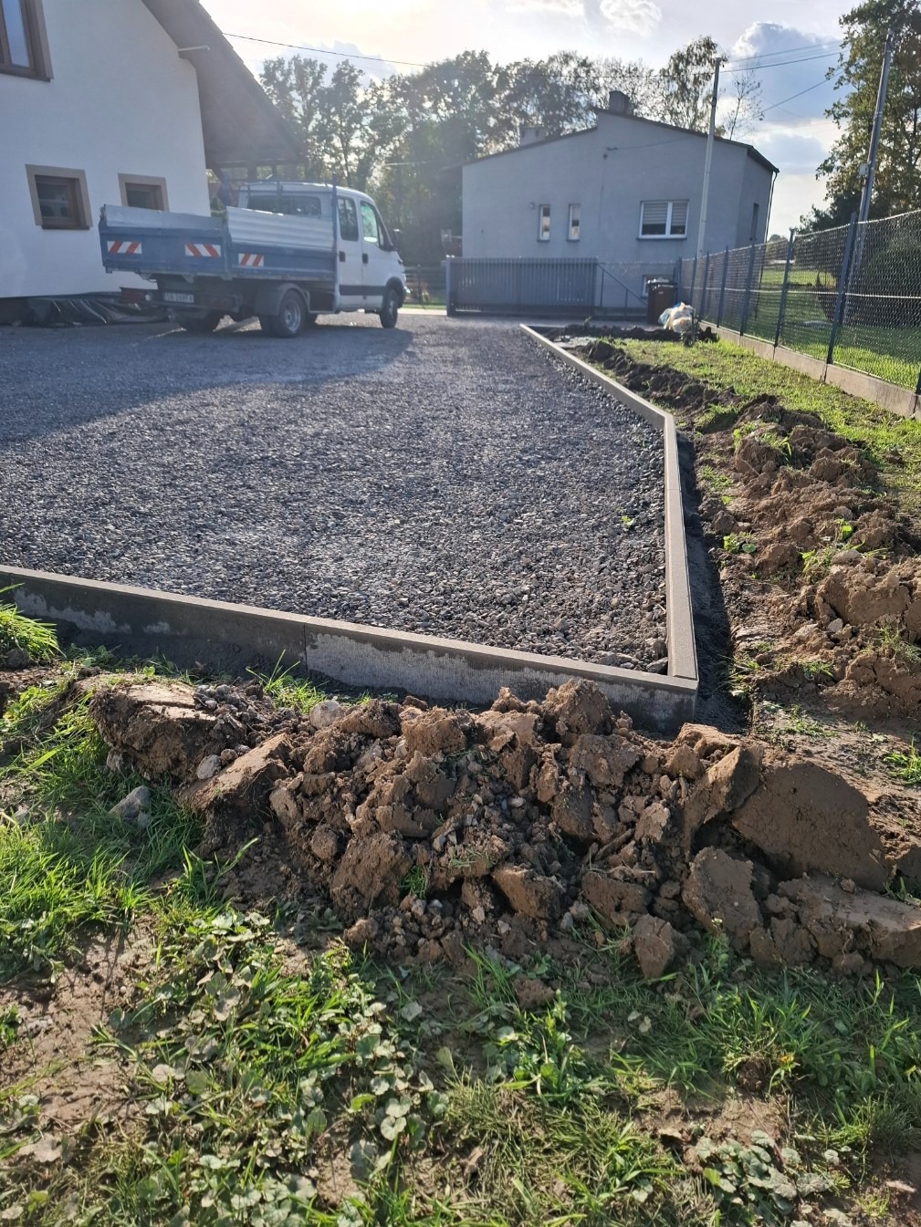 Wyrównany teren z szarą nawierzchnią z kamieni, obrzeża z betonowych krawężników. W tle budynki i samochód dostawczy. Przygotowanie terenu pod budowę.