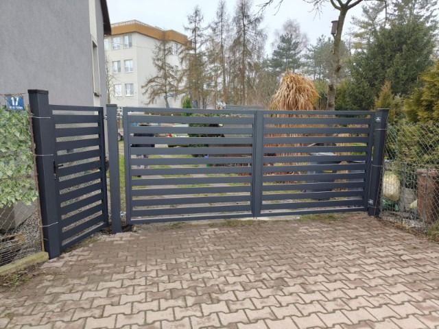 Nowoczesna, grafitowa brama wjazdowa z poziomymi szczeblami, częściowo otwarta furtka, kostka brukowa, zieleń w tle. Minimalistyczny design bramy.