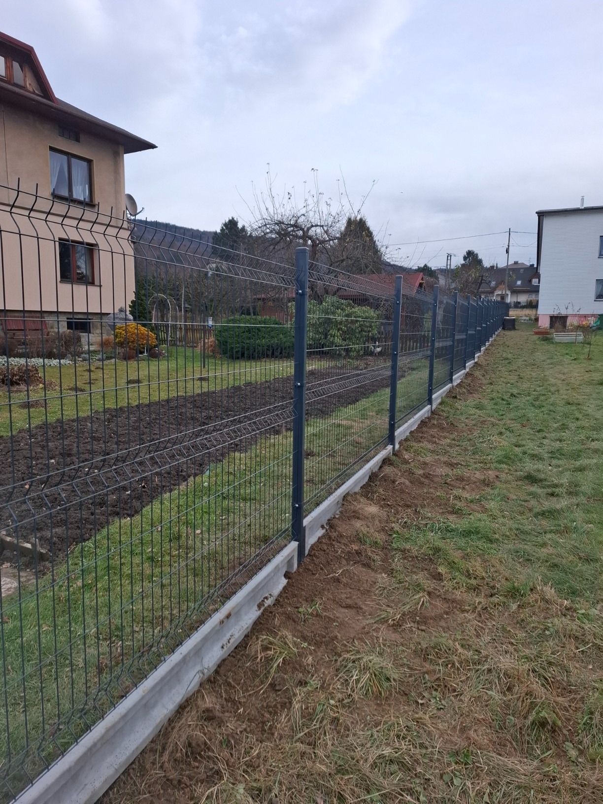 Długi, panelowy płot z podmurówką betonową, ciągnący się wzdłuż granicy działki. Widoczne domy w tle i świeżo przekopana ziemia wzdłuż ogrodzenia.