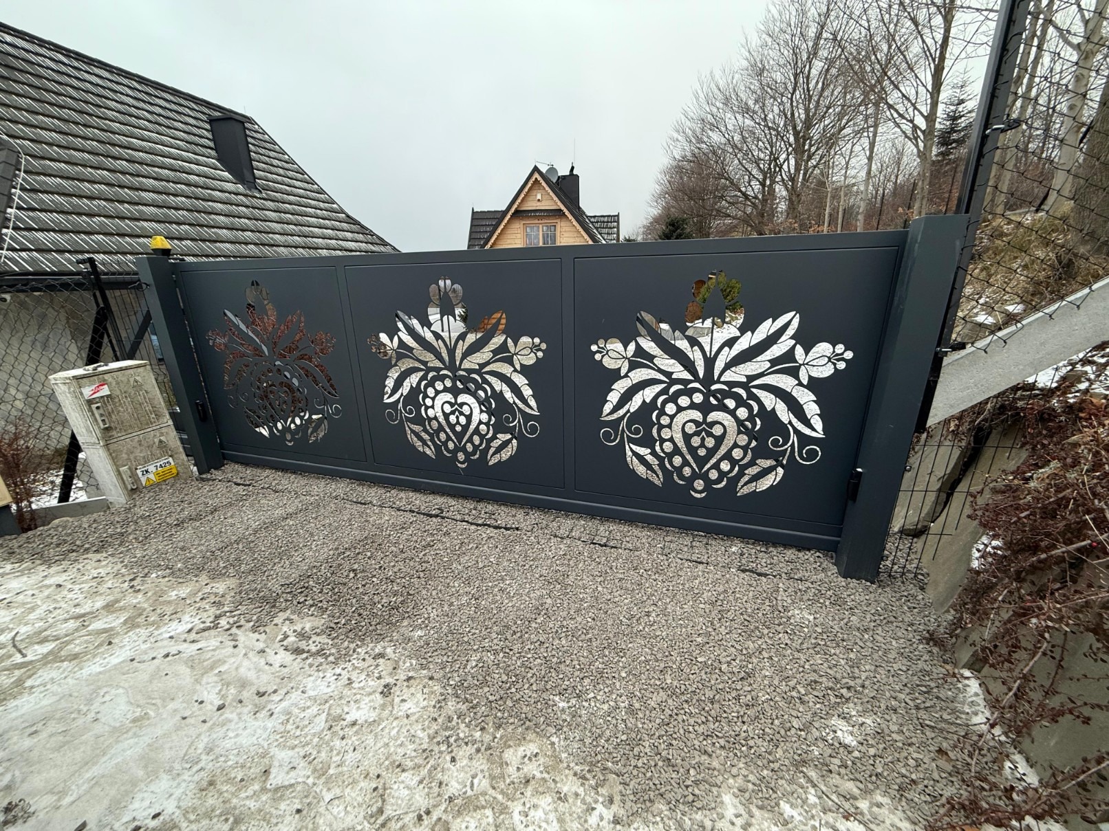 Nowoczesna, przesuwna brama w kolorze grafitowym z ażurowym, folkowym wzorem. Brama zamontowana na posesji z domem w stylu góralskim. Podłoże wysypane drobnym kamieniem.