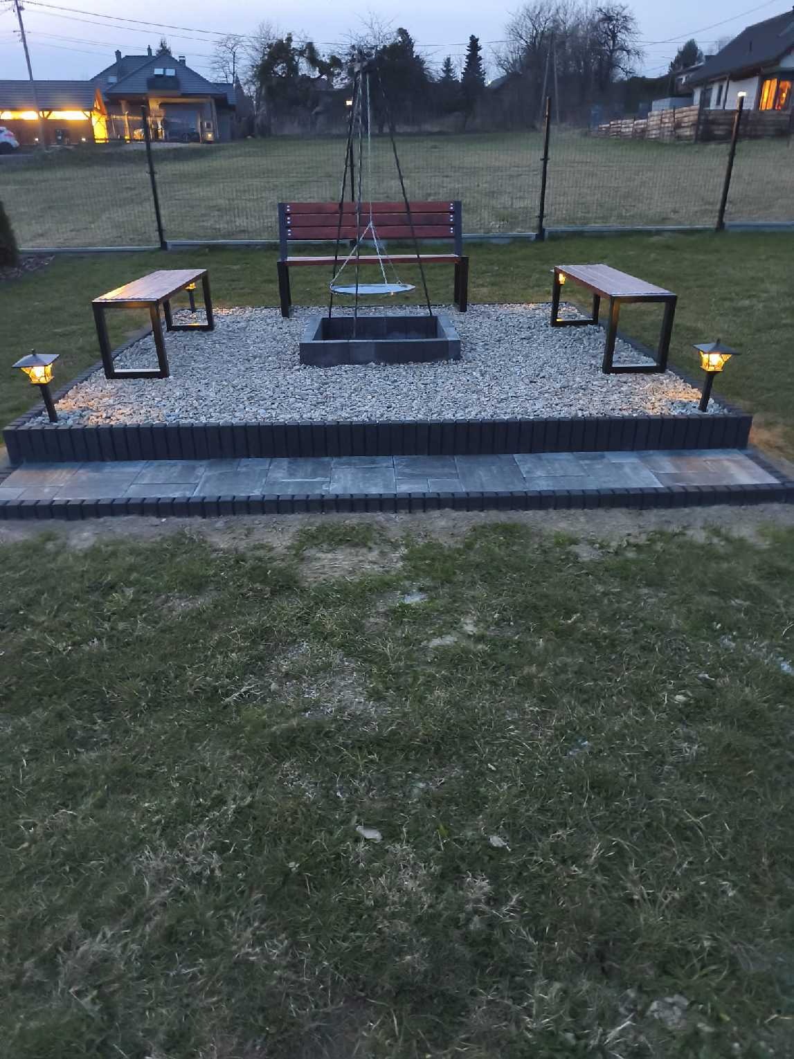 Ogród z miejscem na ognisko: kamienny krąg, ławki z drewna i metalu, oświetlenie. Całość otoczona trawnikiem i ogrodzeniem z siatki.