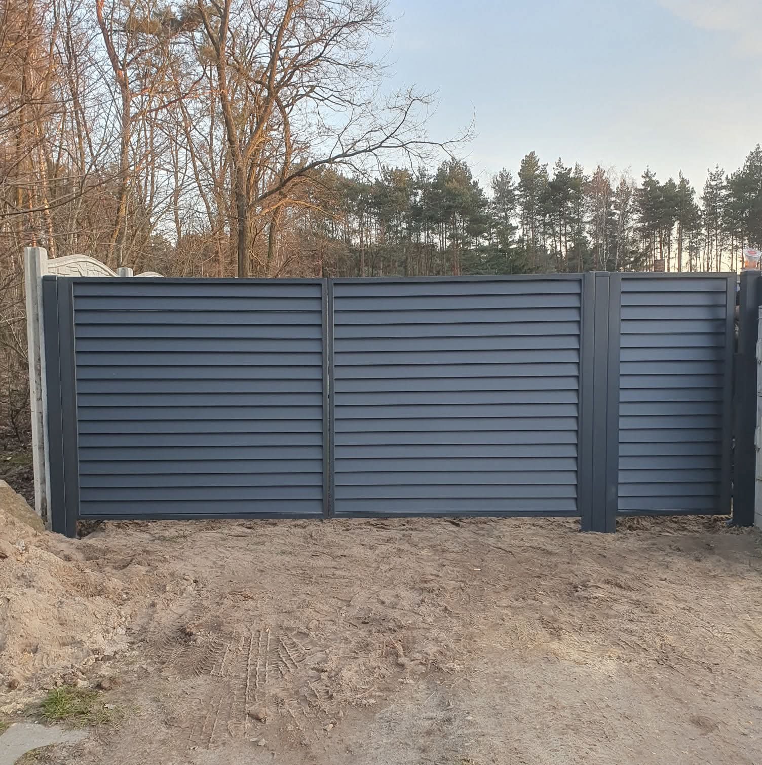 Nowoczesne, poziome ogrodzenie panelowe w kolorze grafitowym, z betonowymi słupkami, na tle drzew i nieba. Widoczne ślady opon na piasku.