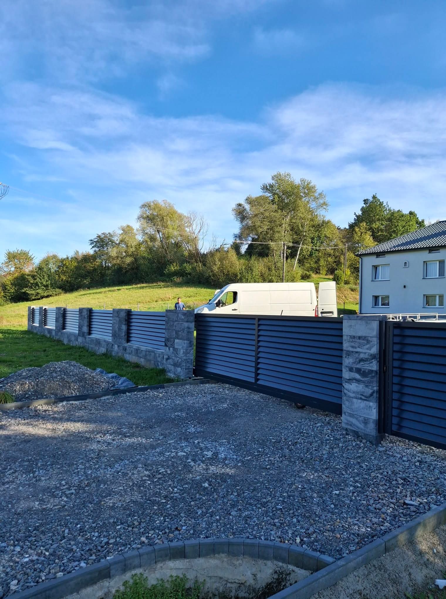 Nowoczesne ogrodzenie panelowe z przesuwną bramą, szare panele i kamienne słupki, na tle zielonego trawnika i błękitnego nieba. Widoczny biały bus.