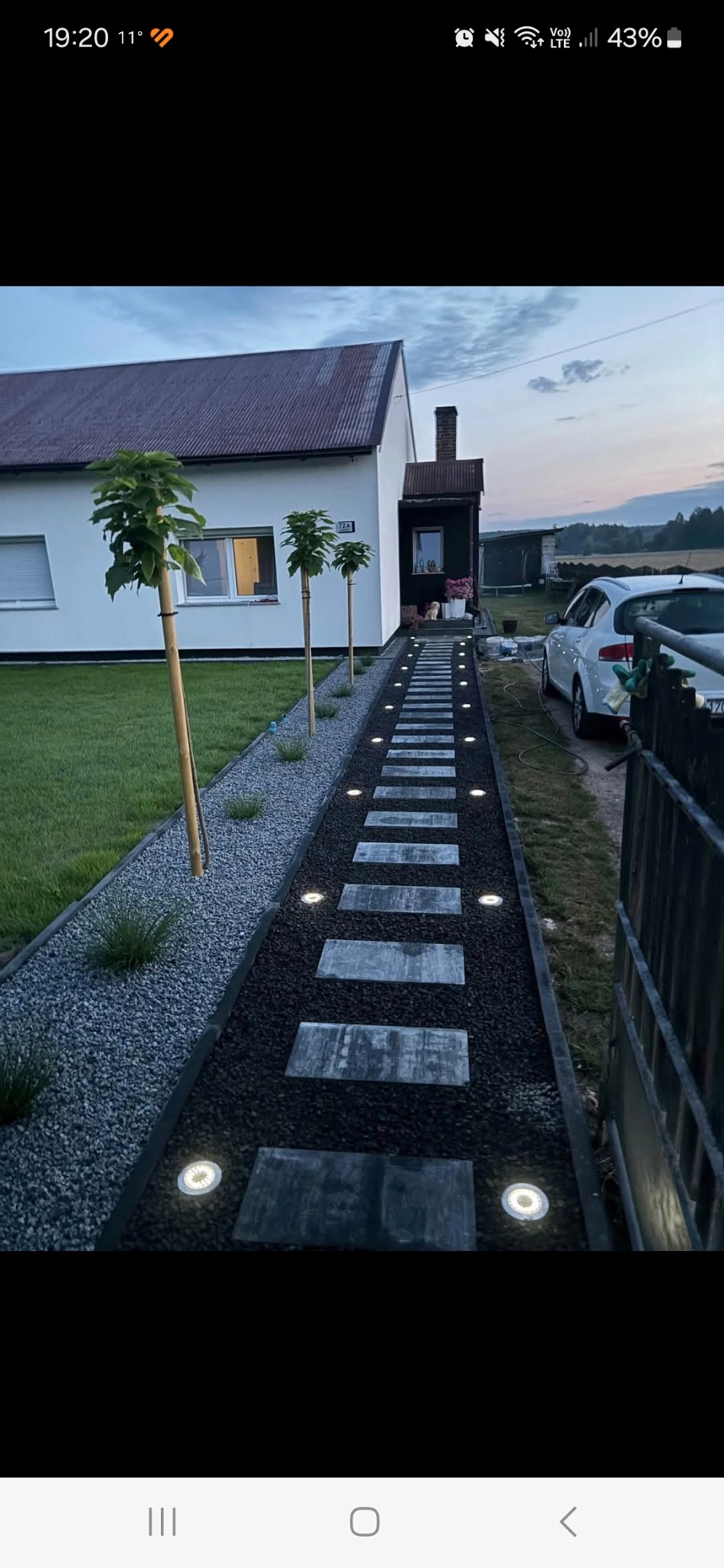 Oświetlona ścieżka z płyt prowadząca do domu, obok trawnik i młode drzewka. Dekoracyjny żwir, lampy wbudowane w ścieżkę. Widok na dom i fragment samochodu.