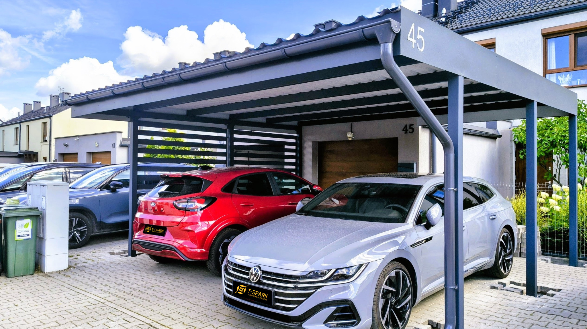 Nowoczesna, szara konstrukcja zadaszenia dla samochodów, z numerem domu '45', na tle błękitnego nieba z chmurami i zaparkowanymi autami.