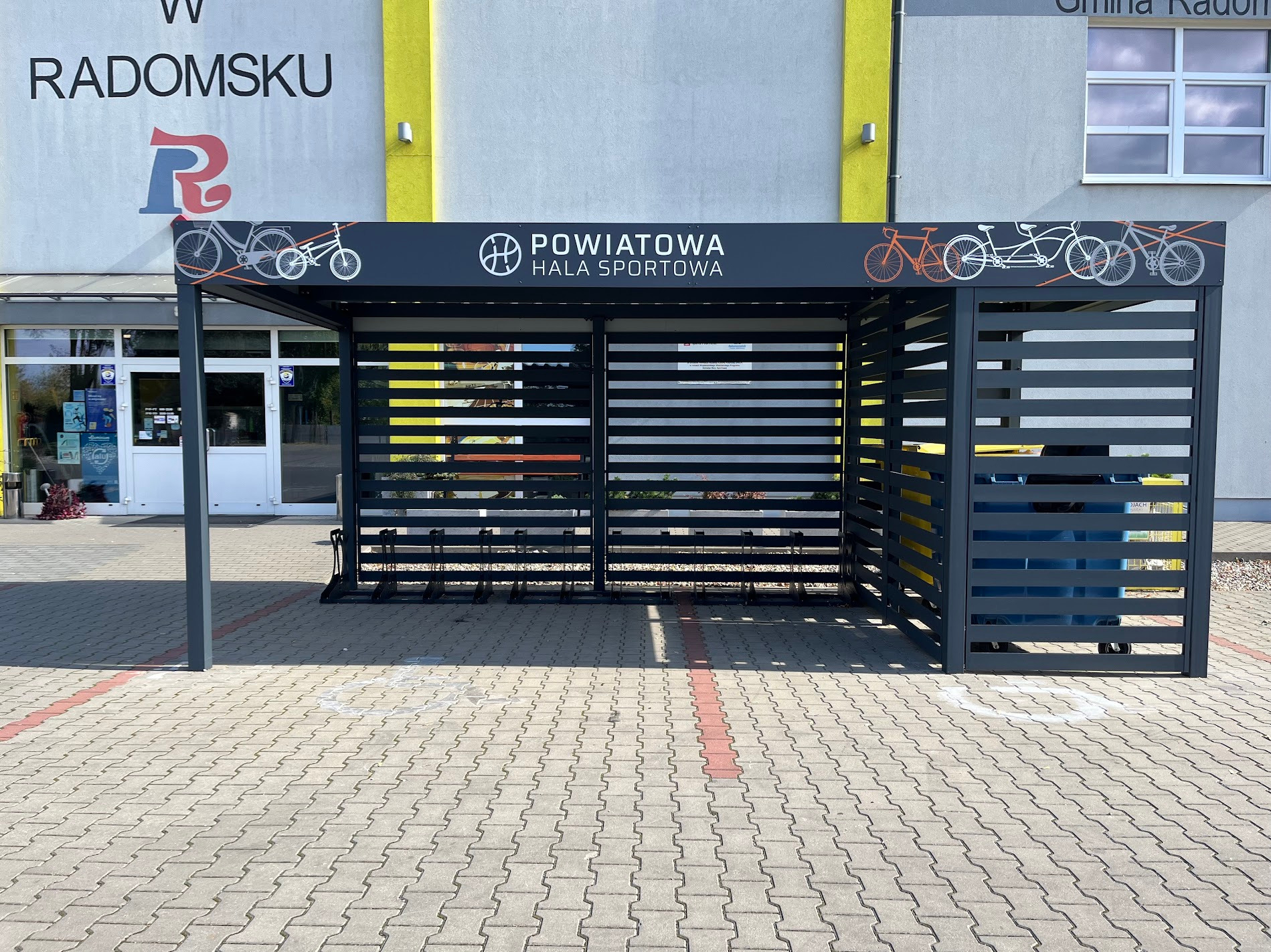Nowoczesna, grafitowa konstrukcja z poziomych listew, z grafiką rowerów, przed budynkiem Powiatowej Hali Sportowej. Widok z zewnątrz, na kostce brukowej.