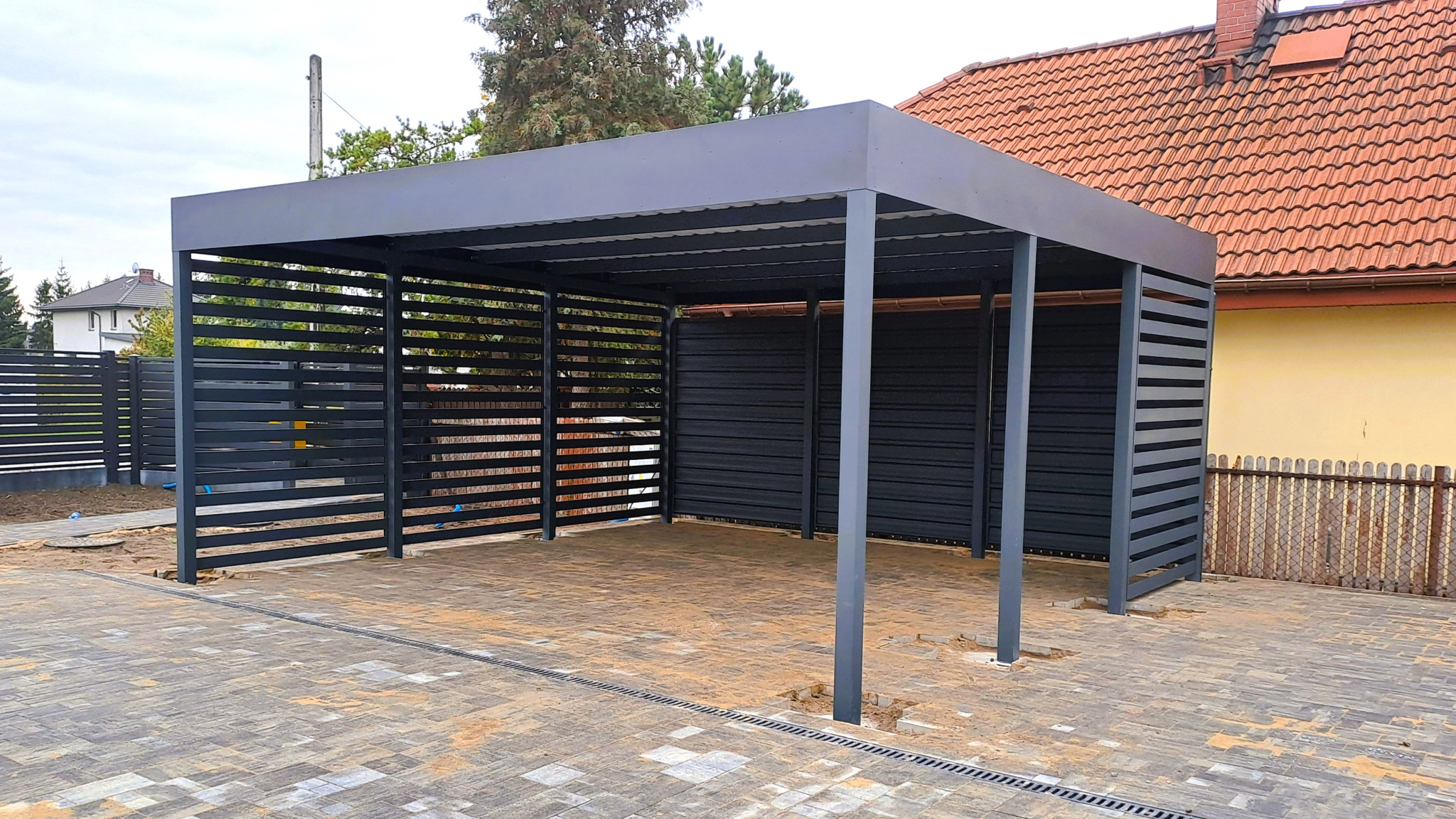 Nowoczesna, grafitowa pergola metalowa z lamelami, na kostce brukowej. Widoczny fragment domu z czerwonym dachem i ogrodzenie. Minimalistyczny design.