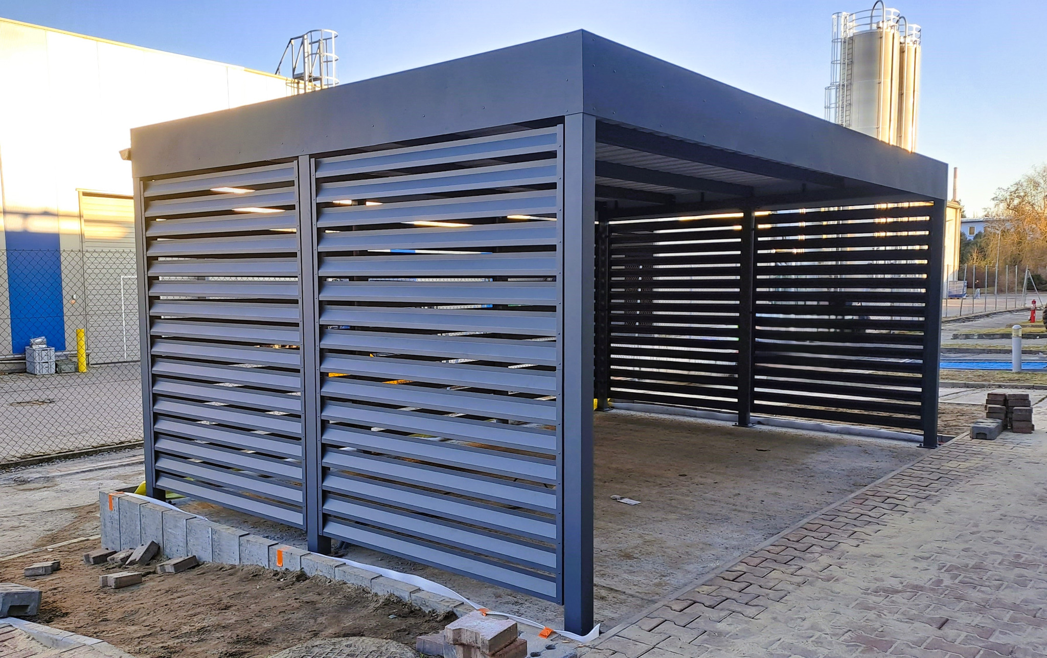 Nowoczesna, szara konstrukcja z metalowych paneli lamelowych, prawdopodobnie zadaszenie tarasu lub wiata, w trakcie montażu, z kostką brukową w tle.