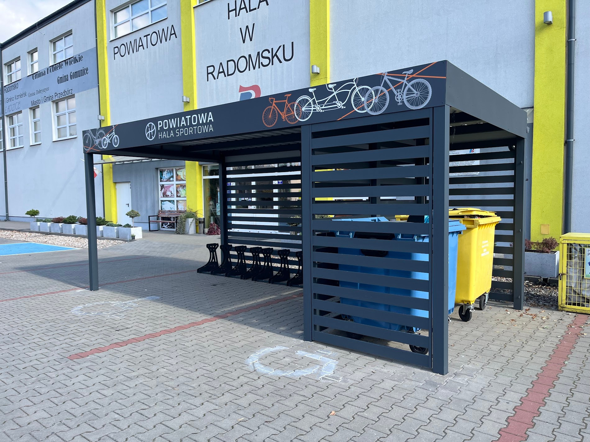 Szara wiata rowerowa z metalowymi lamelami, ozdobiona grafiką rowerów, na tle budynku Powiatowej Hali Sportowej w Radomsku. Widoczne kosze na śmieci.