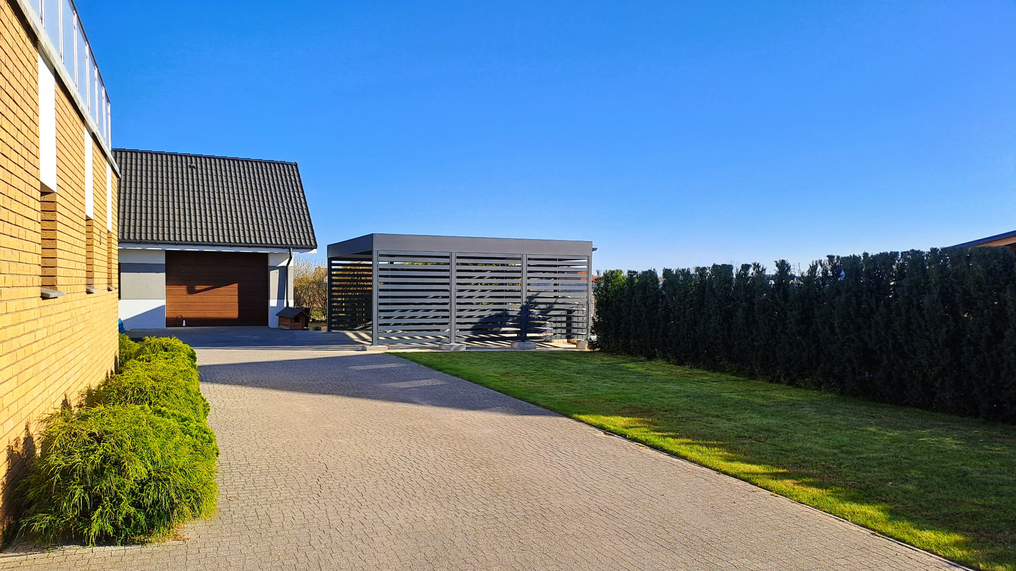 Nowoczesna, szara konstrukcja z poziomych listew, pełniąca funkcję zadaszenia na posesji. Widok z perspektywy chodnika, w tle budynek z garażem i zielony żywopłot.